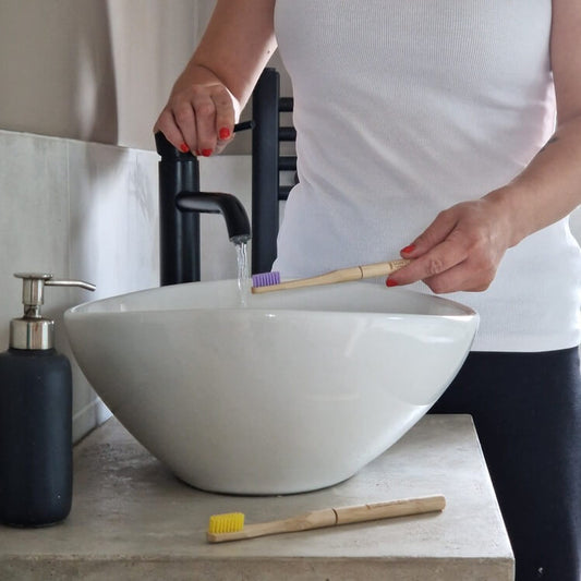 A person holding a bamboo toothbrush over a sink with water running.
