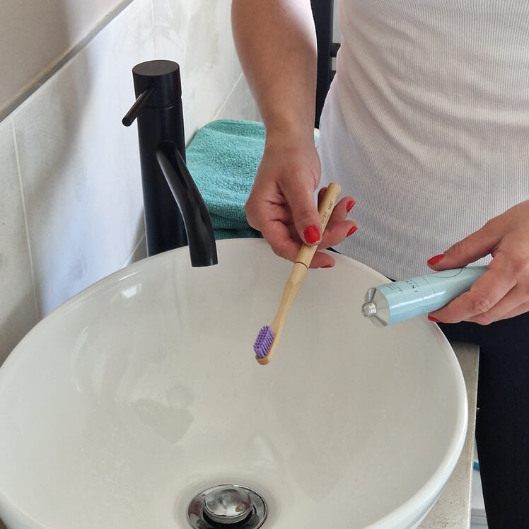 A person holding a bamboo toothbrush with purple bristles and toothpaste over a sink.